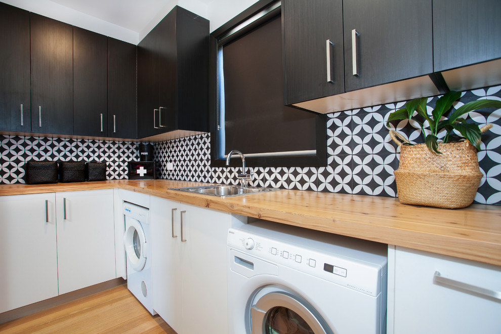 Photo of a contemporary l-shaped dedicated laundry room in Melbourne with a drop-in sink, flat-panel cabinets, black cabinets, wood benchtops, multi-coloured walls, light hardwood floors and beige benchtop.