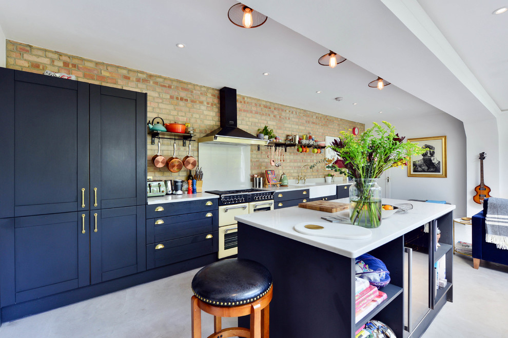 This is an example of an eclectic single-wall open plan kitchen in London with a farmhouse sink, shaker cabinets, brown splashback, brick splashback, coloured appliances, concrete floors, with island, grey floor and blue cabinets.