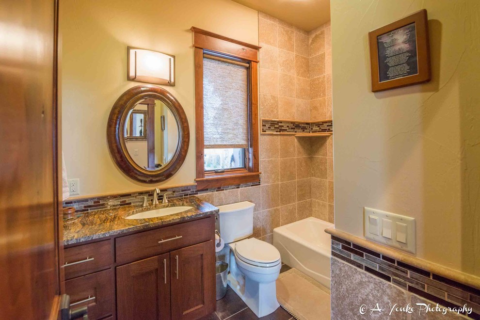 This is an example of a mid-sized country 3/4 bathroom in Other with shaker cabinets, medium wood cabinets, an alcove tub, a shower/bathtub combo, a one-piece toilet, beige tile, brown tile, gray tile, ceramic tile, yellow walls, slate floors, an undermount sink and granite benchtops.