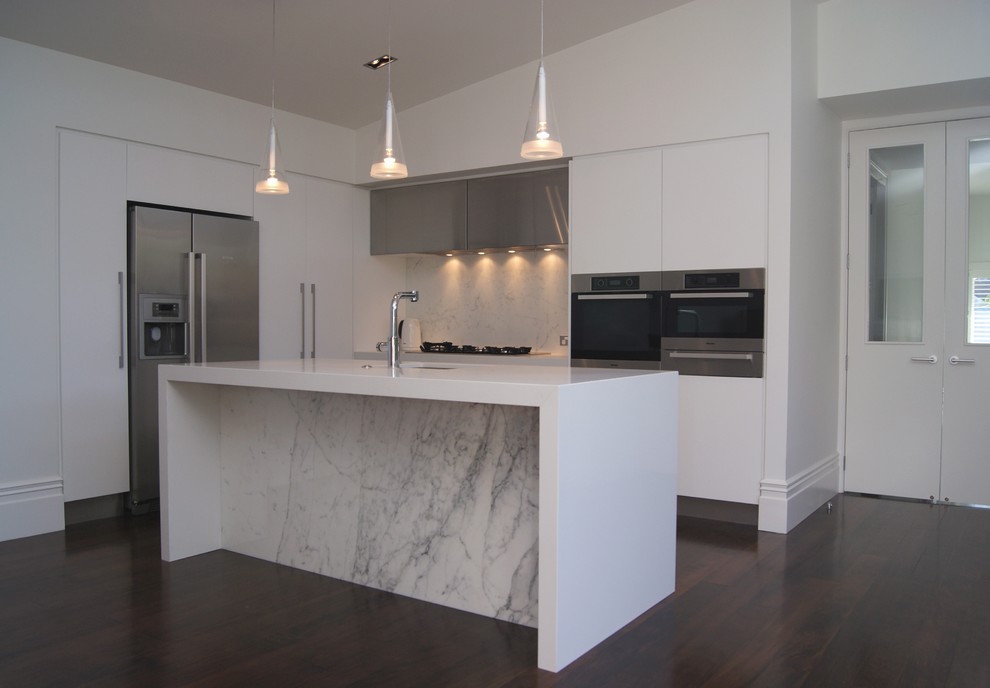 This is an example of a small contemporary l-shaped eat-in kitchen in Auckland with stainless steel appliances, dark hardwood floors, an undermount sink, flat-panel cabinets, white cabinets, quartz benchtops, stone slab splashback and with island.