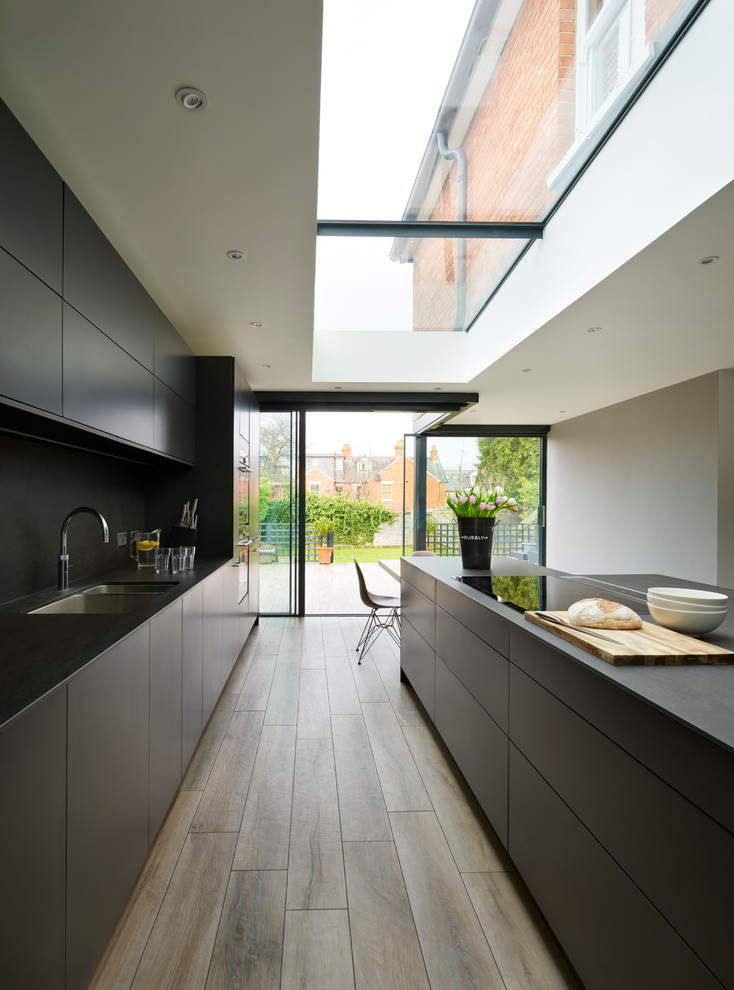 Photo of a modern galley eat-in kitchen in Berkshire with a double-bowl sink, flat-panel cabinets, black cabinets, black splashback, panelled appliances, medium hardwood floors, with island, brown floor and black benchtop.