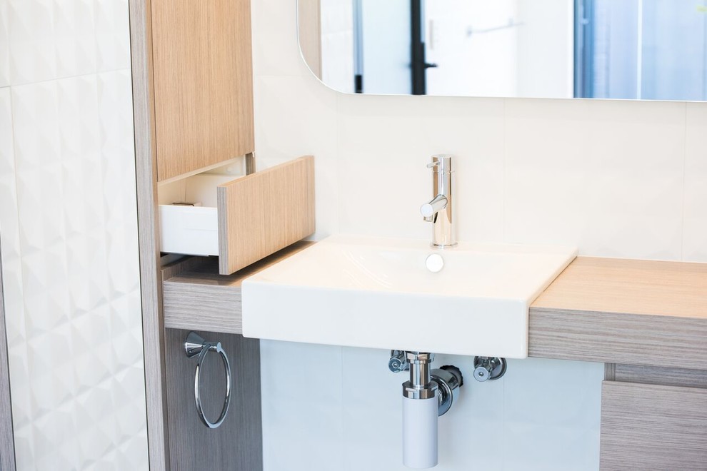 Inspiration for a modern bathroom in Melbourne with flat-panel cabinets, light wood cabinets, a curbless shower, a bidet, gray tile, white tile, porcelain tile, white walls, a wall-mount sink and wood benchtops.