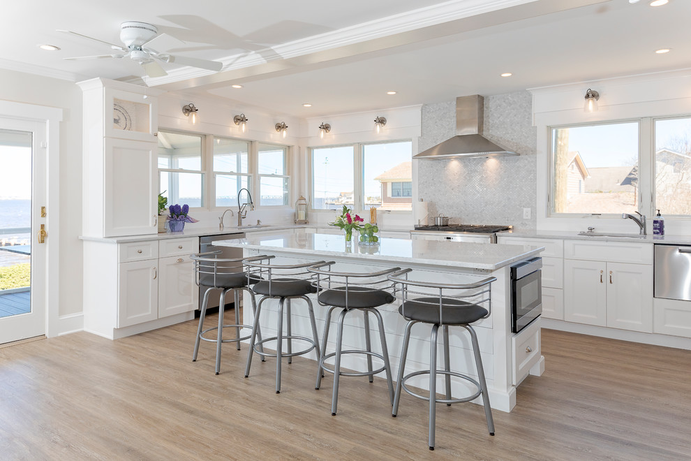Mid-sized beach style u-shaped eat-in kitchen in New York with an undermount sink, shaker cabinets, white cabinets, quartzite benchtops, grey splashback, marble splashback, stainless steel appliances, painted wood floors, with island, brown floor and white benchtop.