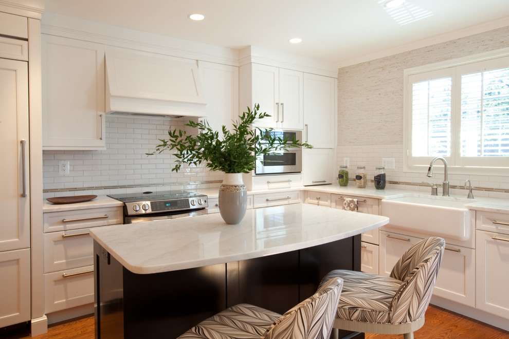 This is an example of a mid-sized transitional l-shaped eat-in kitchen in Baltimore with a farmhouse sink, recessed-panel cabinets, white cabinets, marble benchtops, white splashback, subway tile splashback, stainless steel appliances, medium hardwood floors, with island, brown floor and white benchtop.