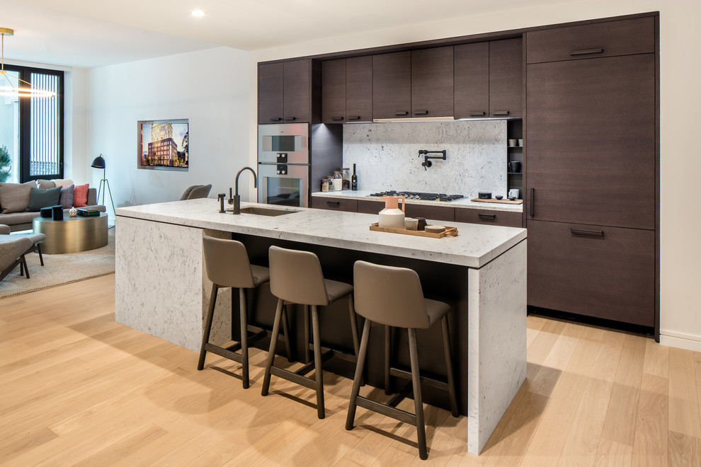 Large contemporary single-wall open plan kitchen in New York with an undermount sink, flat-panel cabinets, dark wood cabinets, marble benchtops, white splashback, marble splashback, stainless steel appliances, light hardwood floors, with island and brown floor.