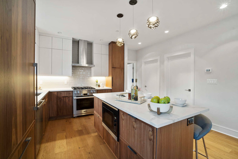 This is an example of a mid-sized modern l-shaped eat-in kitchen in Boston with a drop-in sink, flat-panel cabinets, quartzite benchtops, white splashback, ceramic splashback, stainless steel appliances, medium hardwood floors, with island, brown floor, white benchtop and medium wood cabinets.