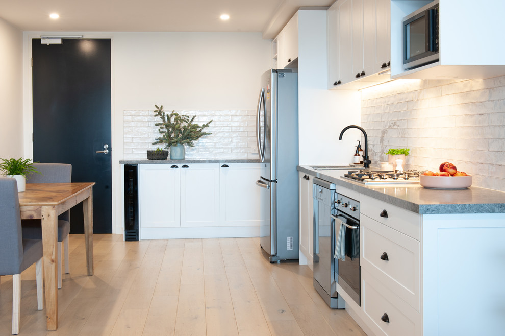 Small modern l-shaped kitchen in Melbourne with a double-bowl sink, recessed-panel cabinets, white cabinets, laminate benchtops, white splashback, subway tile splashback, stainless steel appliances, light hardwood floors, with island and grey benchtop.