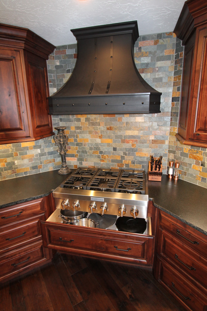 Large mountain style medium tone wood floor and brown floor kitchen photo with raised-panel cabinets, medium tone wood cabinets, granite countertops, multicolored backsplash, stone tile backsplash, paneled appliances, an island and black countertops
