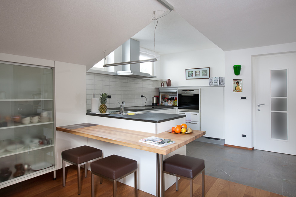 This is an example of a mid-sized contemporary u-shaped eat-in kitchen in Barcelona with white splashback, a peninsula, a drop-in sink, flat-panel cabinets, white cabinets, solid surface benchtops, subway tile splashback, stainless steel appliances and slate floors.
