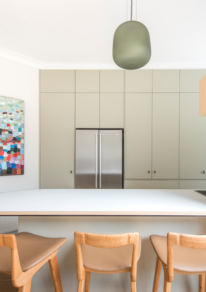 Photo of a contemporary u-shaped eat-in kitchen in Melbourne with a double-bowl sink, flat-panel cabinets, beige cabinets, solid surface benchtops, beige splashback, ceramic splashback, stainless steel appliances, medium hardwood floors, with island, brown floor and white benchtop.