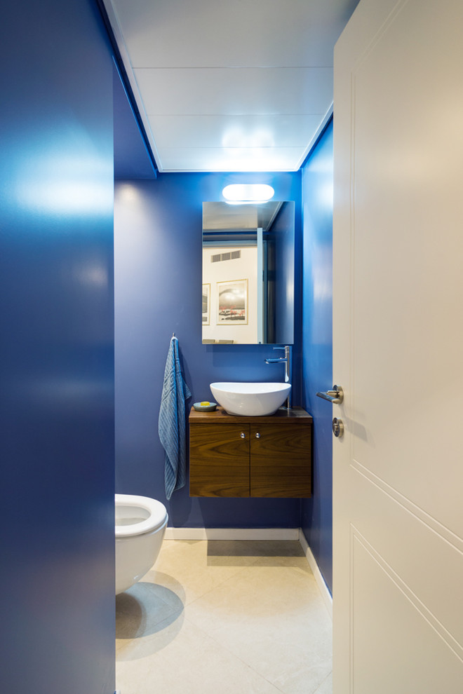 Small contemporary powder room in Tel Aviv with a vessel sink, flat-panel cabinets, wood benchtops, a wall-mount toilet, blue walls, ceramic floors and dark wood cabinets.