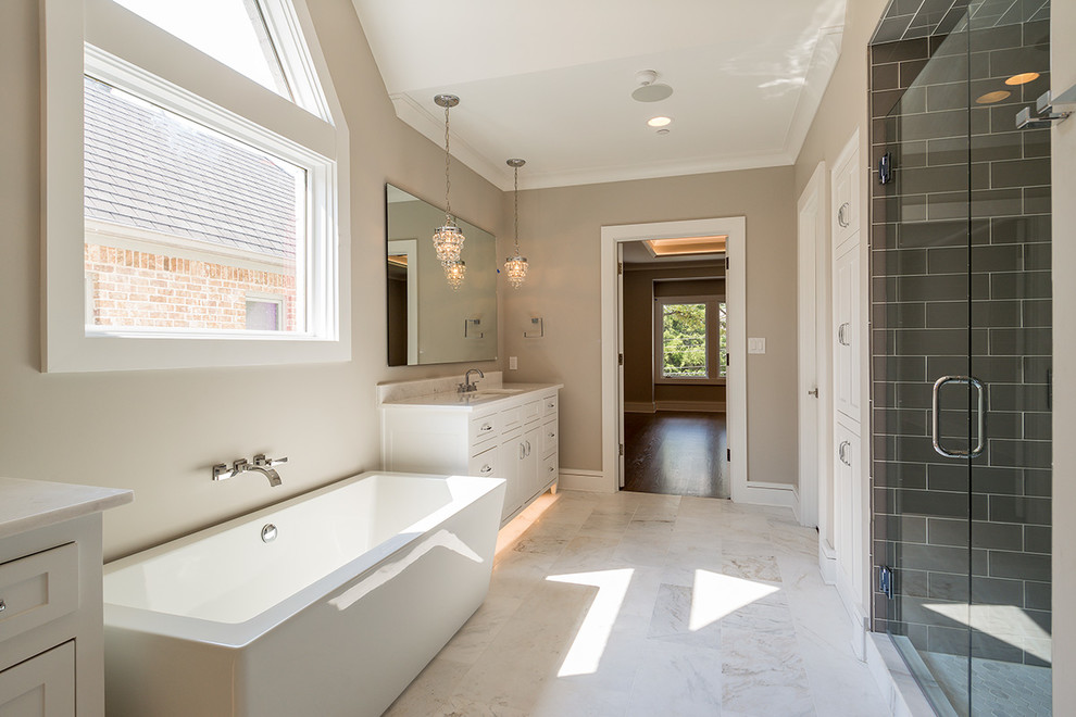 This is an example of a large transitional master bathroom in Dallas with shaker cabinets, white cabinets, a freestanding tub, a double shower, a one-piece toilet, gray tile, glass tile, beige walls, marble floors, an undermount sink, marble benchtops, white floor and a hinged shower door.