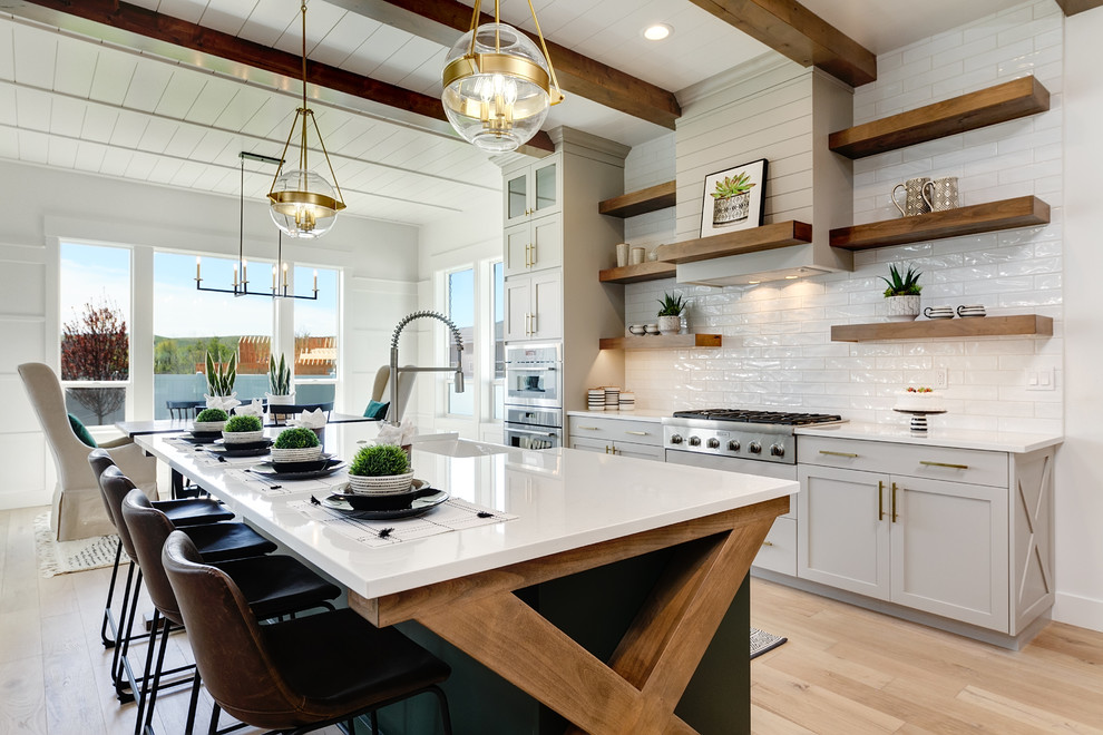 Design ideas for a large country l-shaped open plan kitchen in Boise with a farmhouse sink, shaker cabinets, white cabinets, quartz benchtops, white splashback, subway tile splashback, stainless steel appliances, light hardwood floors, with island, beige floor and white benchtop.