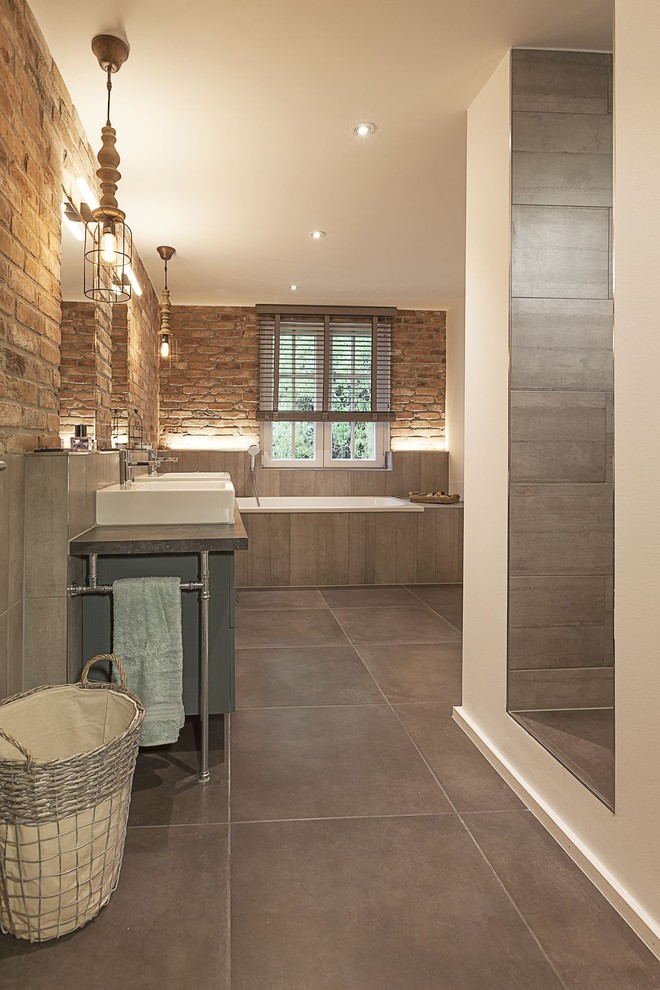 Large industrial bathroom in Dusseldorf with a drop-in tub, brown tile, red tile, cement tiles, stone slab, granite benchtops, grey cabinets, a wall-mount toilet, a vessel sink and grey floor.