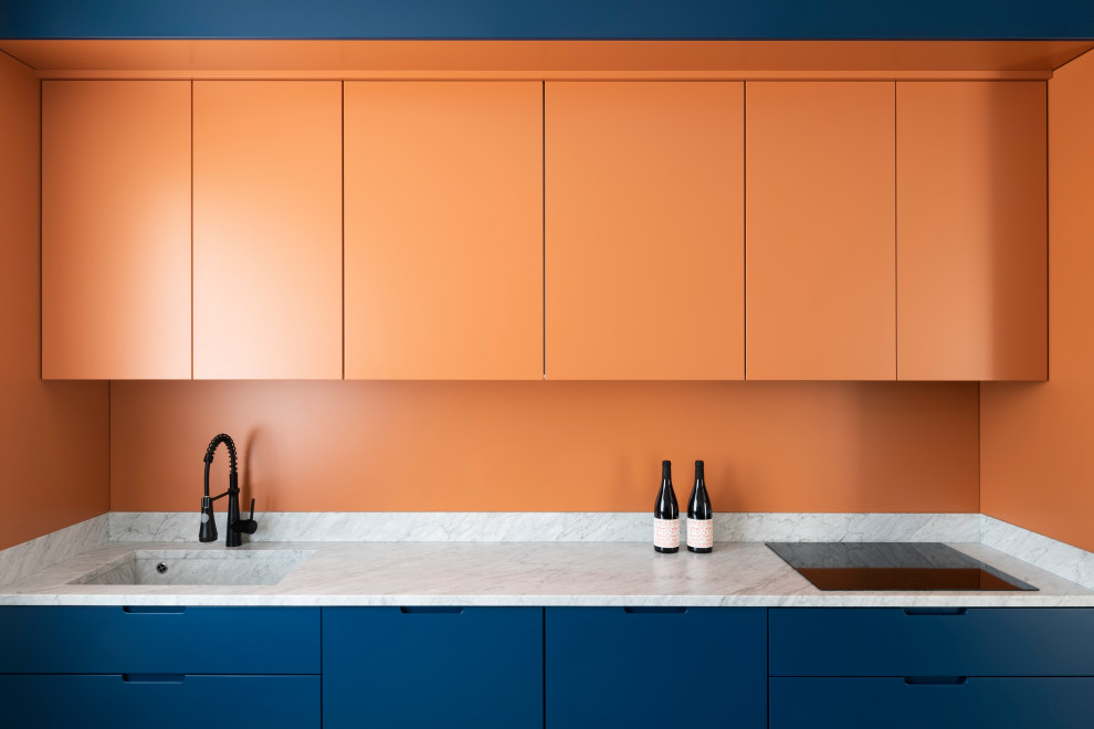 Diseño de cocina lineal actual grande abierta con puertas de armario azules, encimera de mármol, salpicadero naranja, salpicadero de mármol, electrodomésticos de acero inoxidable, suelo de cemento, suelo gris y encimeras blancas