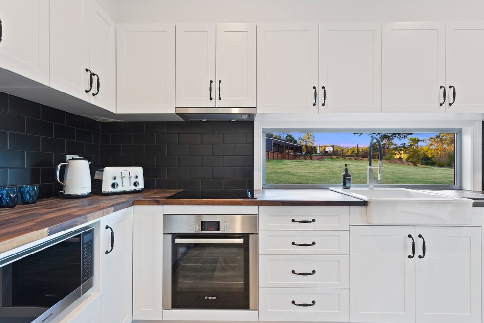 This is an example of a small country kitchen in Sunshine Coast with a farmhouse sink, shaker cabinets, white cabinets, wood benchtops, black splashback, subway tile splashback, stainless steel appliances, travertine floors and multi-coloured floor.