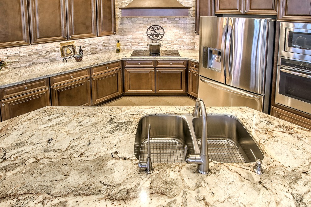 Large transitional l-shaped brown floor open concept kitchen photo in Phoenix with a double-bowl sink, raised-panel cabinets, dark wood cabinets, granite countertops, beige backsplash, stone tile backsplash, stainless steel appliances and an island
