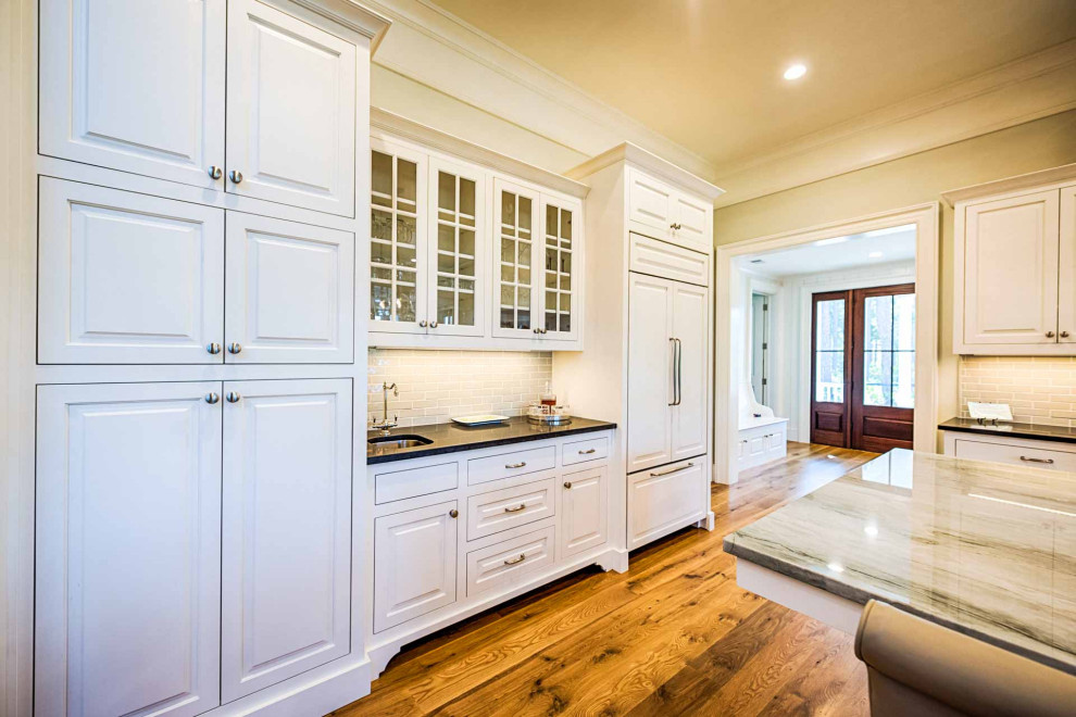 Example of a light wood floor and brown floor eat-in kitchen design with a farmhouse sink, raised-panel cabinets, white cabinets, marble countertops, gray backsplash, ceramic backsplash, stainless steel appliances, an island and multicolored countertops