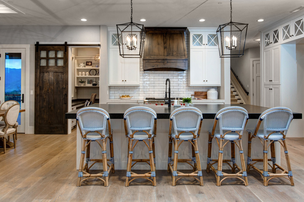 Inspiration for a country l-shaped eat-in kitchen in Salt Lake City with a farmhouse sink, shaker cabinets, white cabinets, white splashback, stainless steel appliances, light hardwood floors and with island.