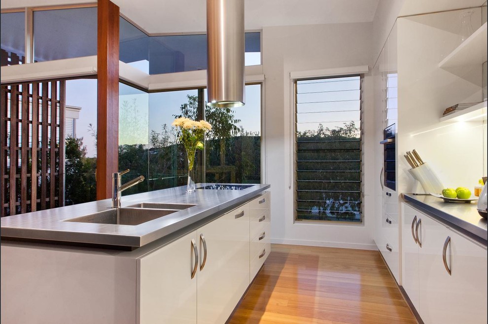 Design ideas for a mid-sized beach style galley open plan kitchen in Sunshine Coast with an undermount sink, flat-panel cabinets, white cabinets, stainless steel benchtops, metallic splashback, glass tile splashback, stainless steel appliances, medium hardwood floors, with island and beige floor.