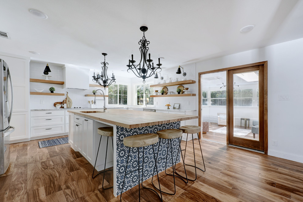 Beach style u-shaped open plan kitchen in Austin with an undermount sink, shaker cabinets, white cabinets, wood benchtops, white splashback, stainless steel appliances, medium hardwood floors, with island and brown floor.