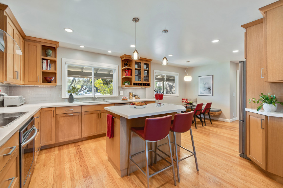 This is an example of a transitional l-shaped eat-in kitchen in San Francisco with an undermount sink, shaker cabinets, medium wood cabinets, grey splashback, glass tile splashback, stainless steel appliances, medium hardwood floors, with island, brown floor and white benchtop.