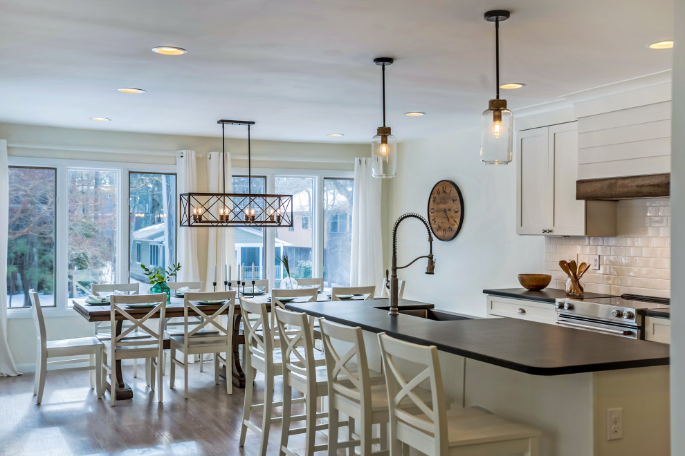 Photo of a large modern l-shaped eat-in kitchen in Other with a farmhouse sink, shaker cabinets, white cabinets, granite benchtops, white splashback, subway tile splashback, stainless steel appliances, light hardwood floors, with island and black benchtop.