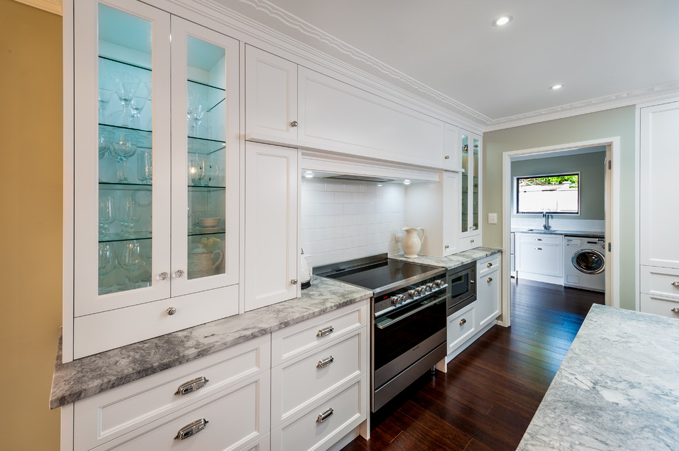 Large traditional eat-in kitchen in Auckland with a farmhouse sink, recessed-panel cabinets, white cabinets, granite benchtops, white splashback, subway tile splashback, stainless steel appliances, medium hardwood floors, with island and brown floor.
