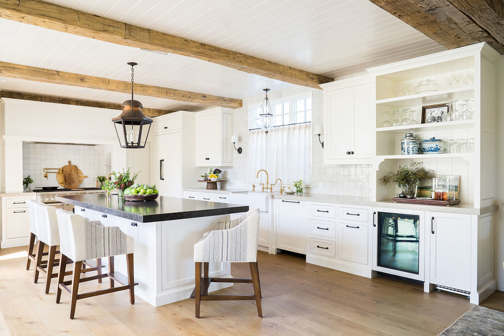 This is an example of a beach style kitchen in Orange County with a farmhouse sink, shaker cabinets, white cabinets, white splashback, panelled appliances, light hardwood floors, with island, beige floor and white benchtop.