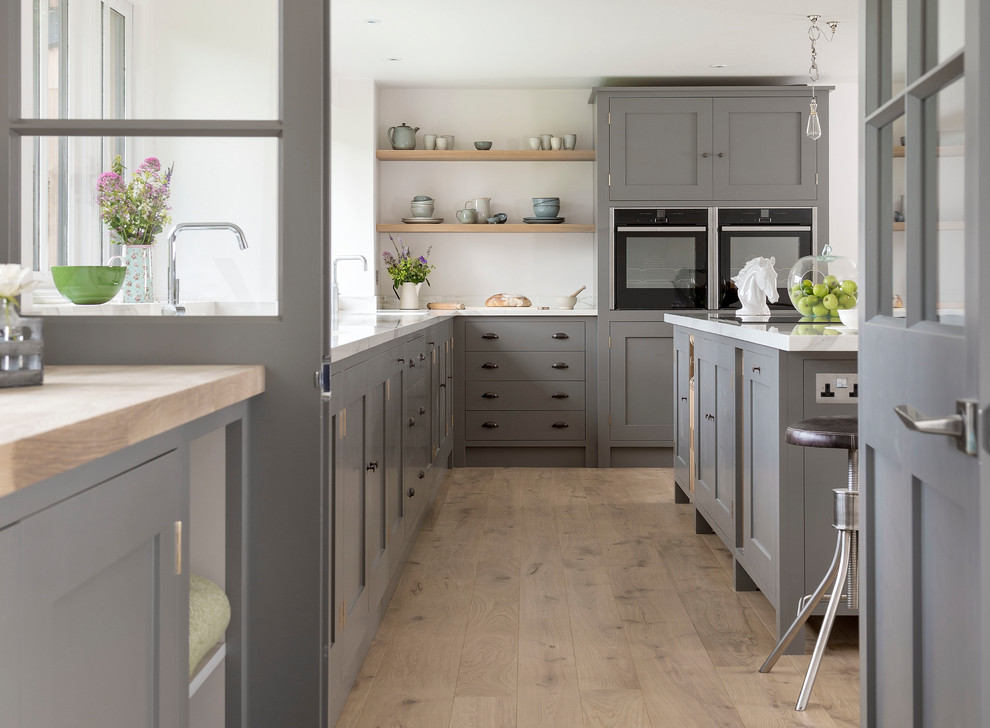 Photo of a mid-sized transitional l-shaped kitchen in Gloucestershire with shaker cabinets, grey cabinets, light hardwood floors, with island, brown floor and white benchtop.