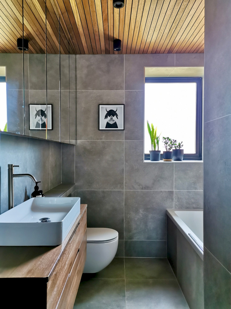 Inredning av ett modernt litet badrum, med släta luckor, bruna skåp, ett platsbyggt badkar, en dusch/badkar-kombination, en vägghängd toalettstol, grå kakel, porslinskakel, grå väggar, klinkergolv i porslin och grått golv