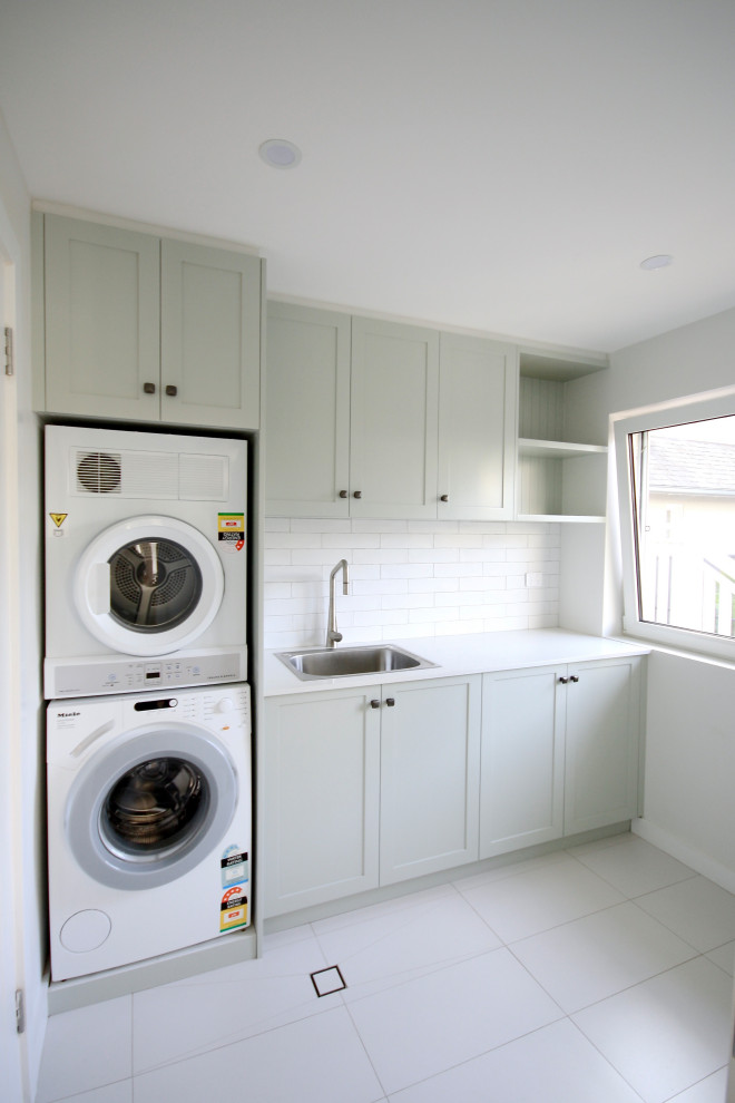 This is an example of a beach style single-wall dedicated laundry room in Sydney with a drop-in sink, shaker cabinets, green cabinets, quartz benchtops, white splashback, subway tile splashback, white walls, ceramic floors, a stacked washer and dryer, white floor and white benchtop.
