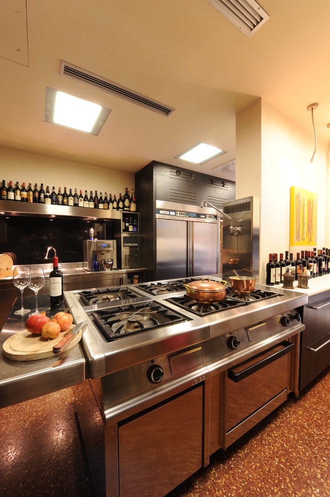 Foto på ett mellanstort funkis kök, med en integrerad diskho, släta luckor, skåp i rostfritt stål, bänkskiva i rostfritt stål, svart stänkskydd, glaspanel som stänkskydd, rostfria vitvaror, terrazzogolv och en köksö