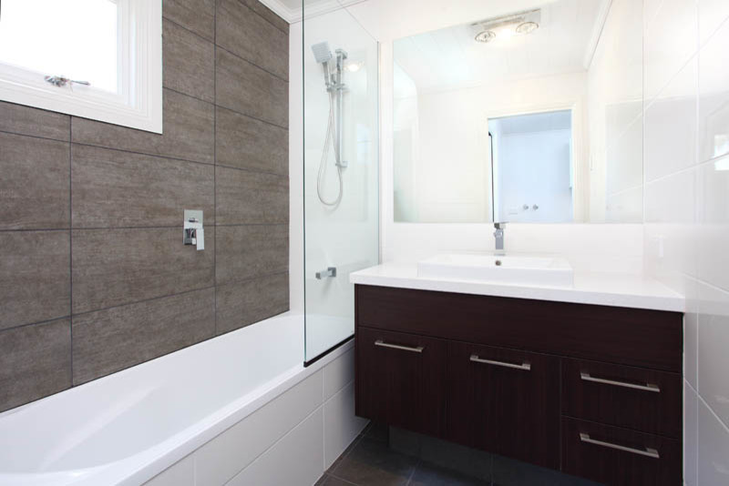 This is an example of a small traditional 3/4 bathroom in Brisbane with flat-panel cabinets, dark wood cabinets, a drop-in tub, a shower/bathtub combo, a one-piece toilet, gray tile, ceramic tile, white walls, ceramic floors, a drop-in sink and engineered quartz benchtops.