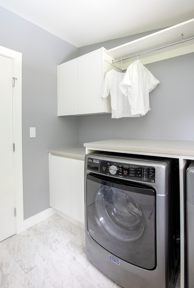 Design ideas for a mid-sized contemporary galley utility room in Vancouver with flat-panel cabinets, white cabinets, grey walls, porcelain floors, a side-by-side washer and dryer, white floor and grey benchtop.