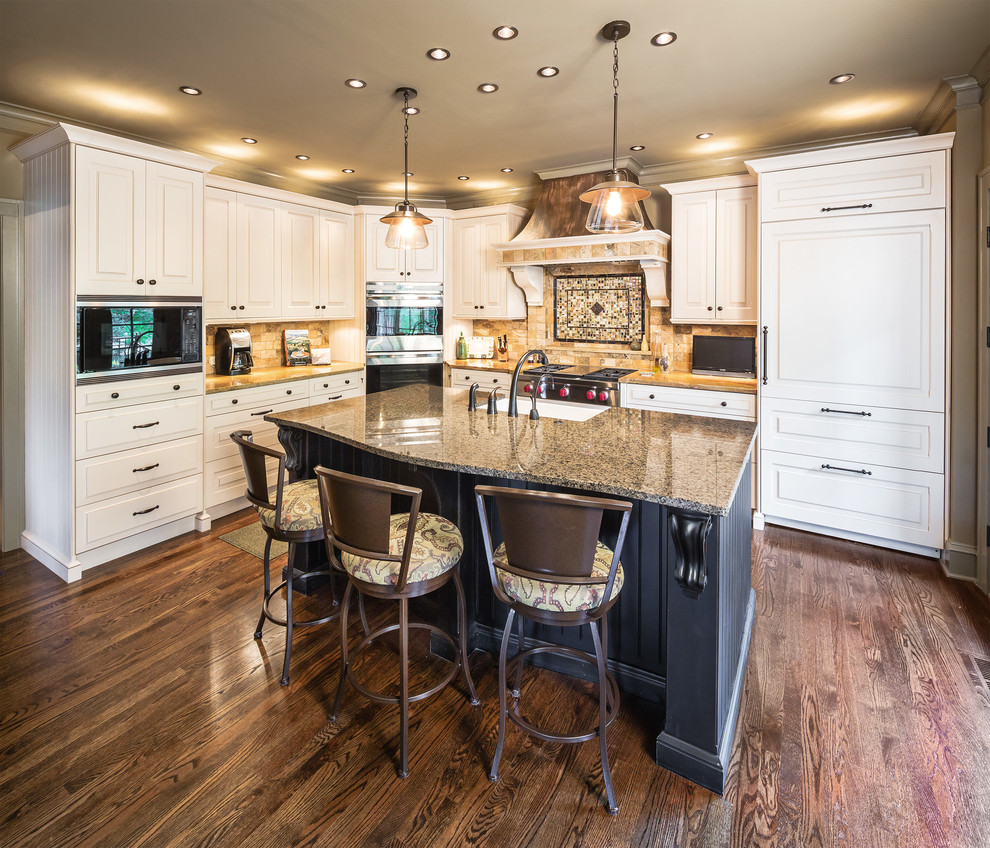 This is an example of a mid-sized traditional l-shaped kitchen in Kansas City with granite benchtops, beige splashback, dark hardwood floors, brown floor, a farmhouse sink, raised-panel cabinets, white cabinets, travertine splashback, panelled appliances and with island.