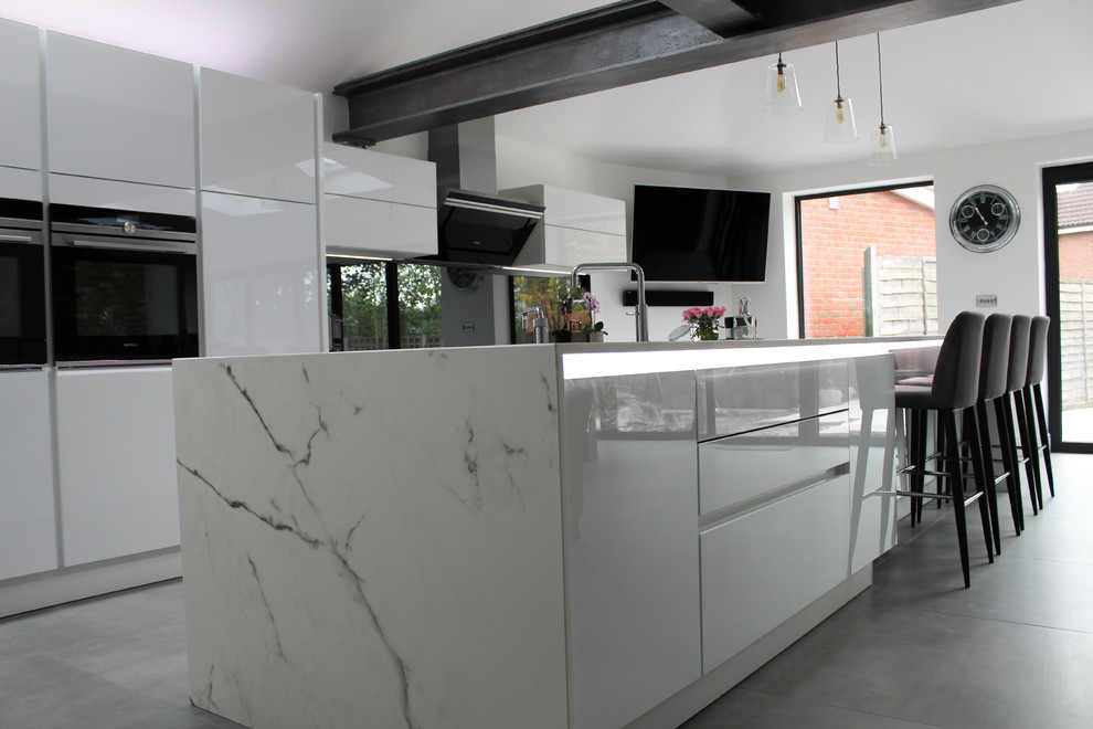 This is an example of a large contemporary eat-in kitchen in Essex with glass-front cabinets, white cabinets, grey splashback, glass sheet splashback, black appliances and with island.