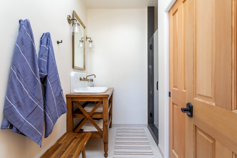 Photo of a mid-sized country bathroom in Santa Barbara with open cabinets, medium wood cabinets, a one-piece toilet, beige tile, porcelain tile, white walls, porcelain floors, wood benchtops, beige floor, a hinged shower door, brown benchtops, an enclosed toilet, a single vanity and a freestanding vanity.