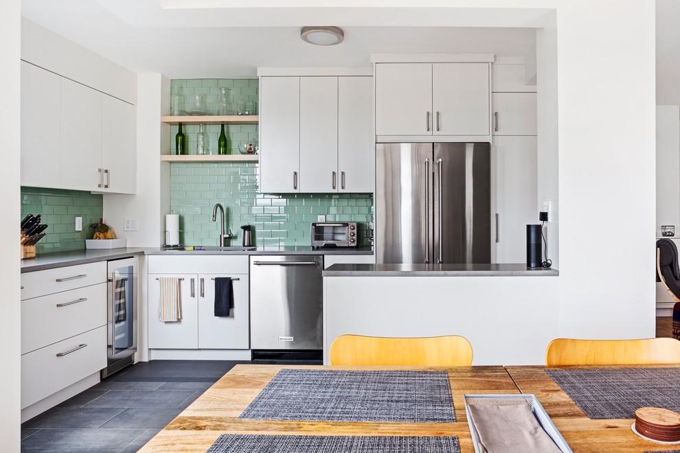 Mid-sized contemporary l-shaped open plan kitchen in New York with an undermount sink, flat-panel cabinets, white cabinets, green splashback, subway tile splashback, stainless steel appliances, a peninsula, grey floor and grey benchtop.