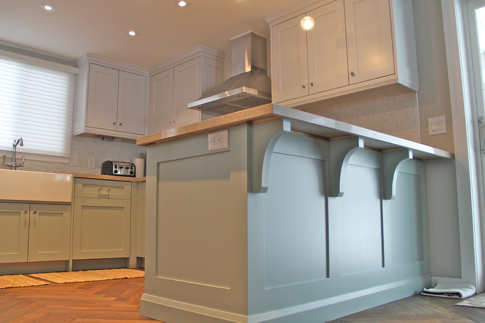 This is an example of a small beach style u-shaped eat-in kitchen in Toronto with a farmhouse sink, shaker cabinets, green cabinets, quartz benchtops, ceramic splashback, stainless steel appliances and light hardwood floors.