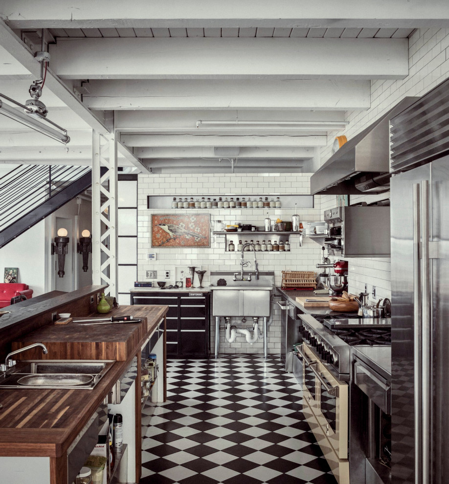 Inspiration for an industrial l-shaped kitchen in Portland Maine with a drop-in sink, flat-panel cabinets, black cabinets, wood benchtops, white splashback, subway tile splashback, stainless steel appliances, with island, multi-coloured floor, brown benchtop and exposed beam.