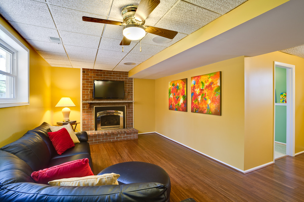 Photo of a large contemporary fully buried basement in Baltimore with yellow walls, dark hardwood floors, a standard fireplace, a brick fireplace surround and brown floor.