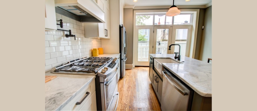 Mid-sized trendy l-shaped medium tone wood floor eat-in kitchen photo in Baltimore with a farmhouse sink, shaker cabinets, white cabinets, marble countertops, white backsplash, subway tile backsplash, stainless steel appliances and an island