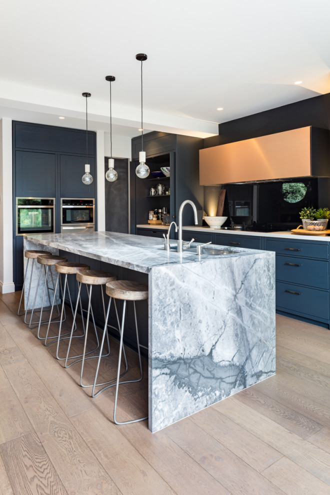 Mid-sized contemporary galley eat-in kitchen in London with a drop-in sink, flat-panel cabinets, blue cabinets, marble benchtops, blue splashback, glass sheet splashback, black appliances, medium hardwood floors, with island, brown floor and grey benchtop.