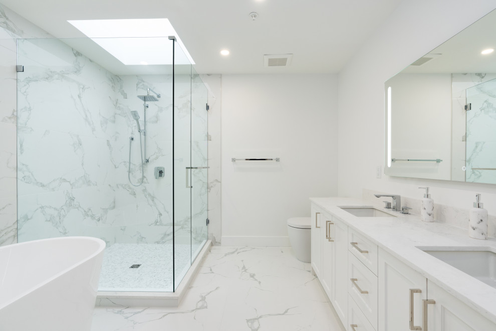 Photo of a large modern master bathroom in Vancouver with shaker cabinets, white cabinets, a freestanding tub, a corner shower, a one-piece toilet, white tile, porcelain tile, white walls, porcelain floors, an undermount sink, engineered quartz benchtops, white floor, a hinged shower door, white benchtops, a double vanity and a built-in vanity.