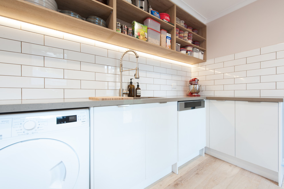 Photo of a mid-sized modern galley utility room in Melbourne with a double-bowl sink, open cabinets, white cabinets, solid surface benchtops, white splashback, subway tile splashback, white walls, light hardwood floors, an integrated washer and dryer and grey benchtop.