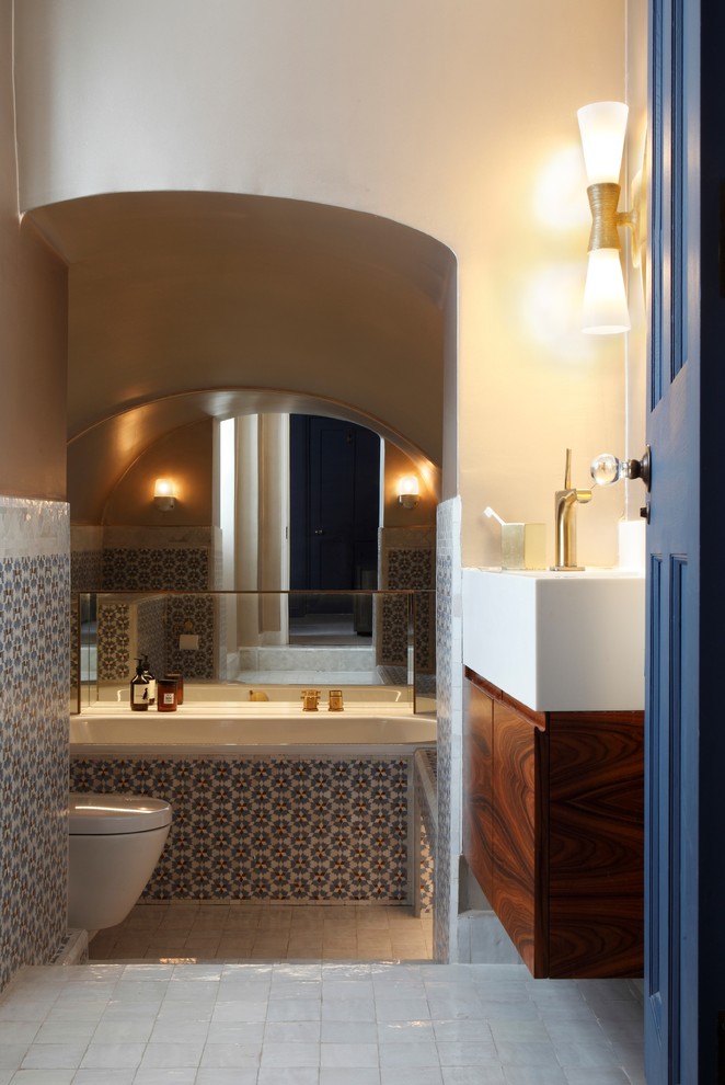 Photo of an eclectic master bathroom in London with flat-panel cabinets, medium wood cabinets, a drop-in tub, multi-coloured tile, mosaic tile, ceramic floors, a wall-mount sink and white floor.