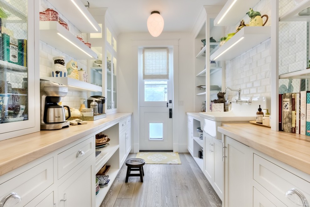 This is an example of a large country galley kitchen pantry in Orange County with a farmhouse sink, recessed-panel cabinets, white cabinets, wood benchtops, white splashback, subway tile splashback and light hardwood floors.