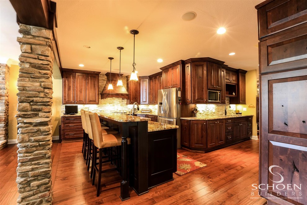 Transitional u-shaped medium tone wood floor kitchen photo with an undermount sink, raised-panel cabinets, dark wood cabinets, granite countertops, beige backsplash, stone tile backsplash, stainless steel appliances and an island