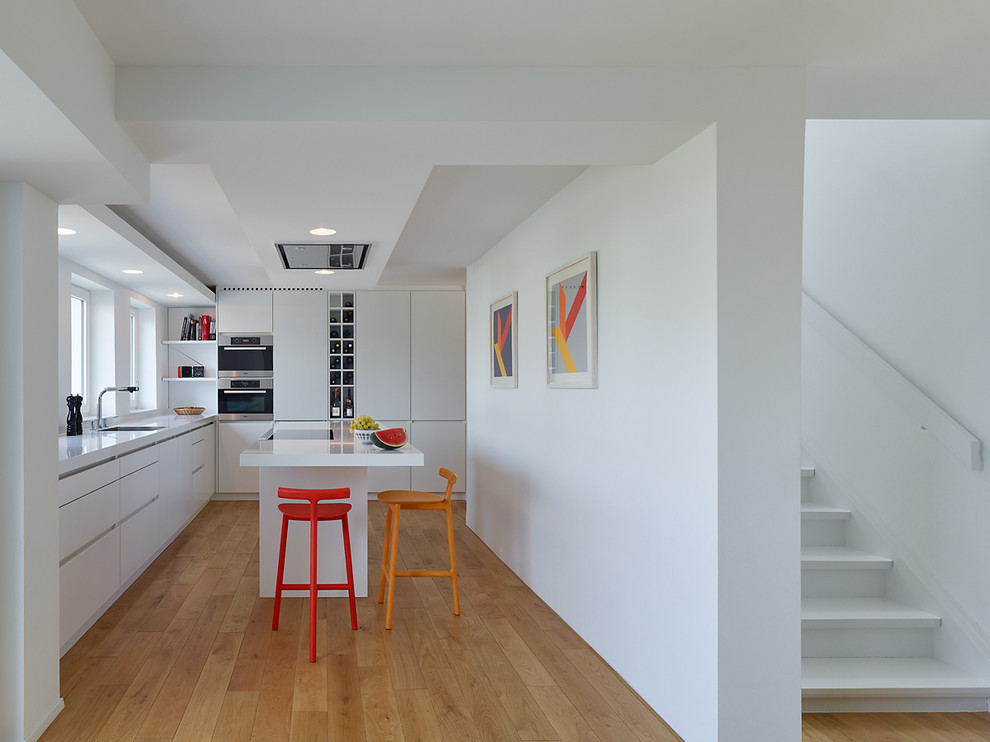 This is an example of a mid-sized contemporary l-shaped open plan kitchen in Stuttgart with flat-panel cabinets, white cabinets, white splashback, stainless steel appliances, medium hardwood floors, with island and a drop-in sink.
